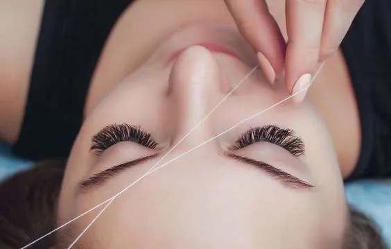 beautician is doing threading on woman's face skin to remove unwanted hairs